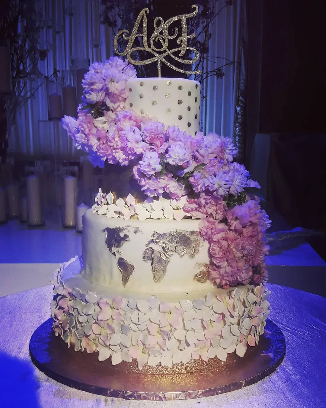 A dramatic tiered wedding cake by AMR Catering decorated with flowers and lit in soft purple light.
