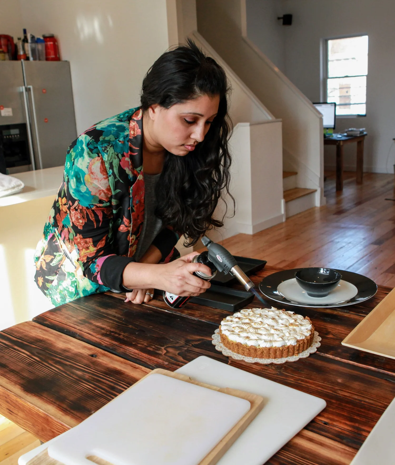 Chef Aarti in a floral jacket gently torching the meringue topping of a tart in her kitchen.